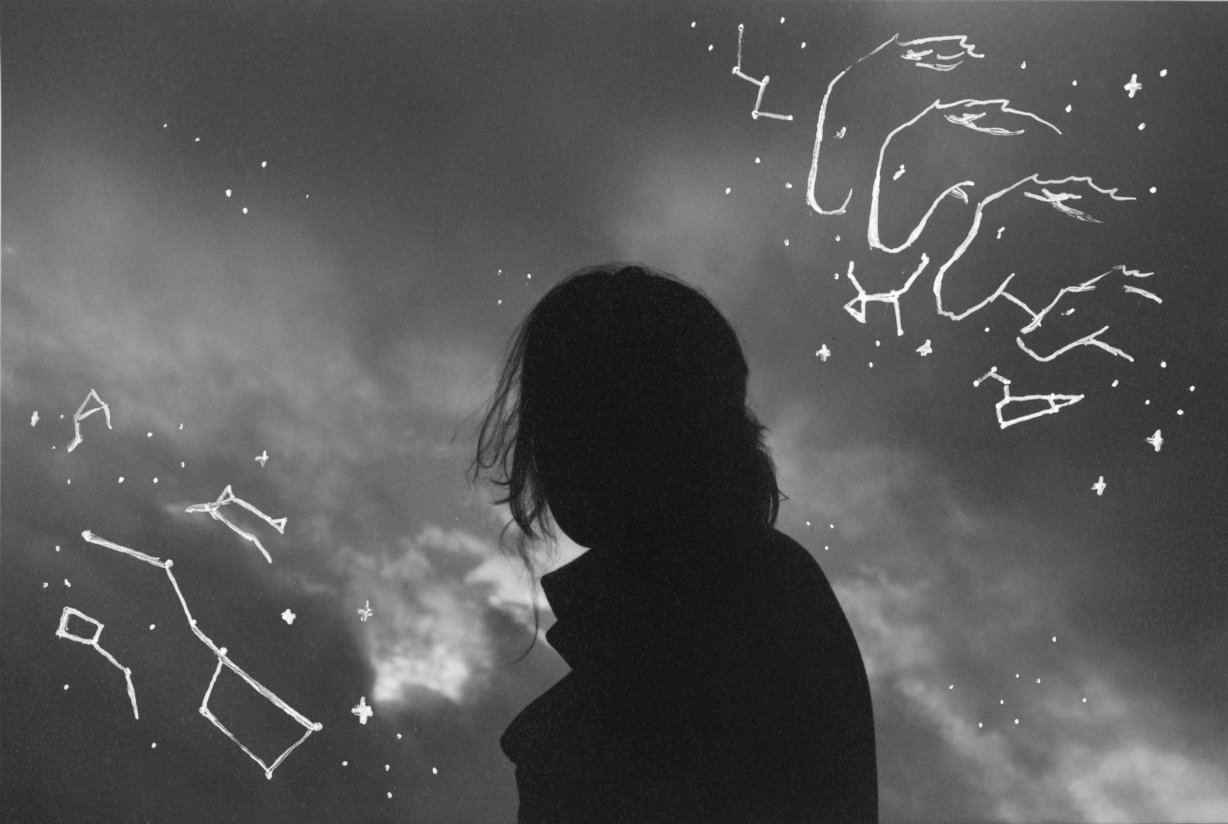 Black and white gelatin silver print of a black silhoette of a woman against a cloudy grey sky. In the sky there are white line drawings of constellations and cave paintings of horses